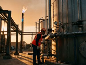 Arbeiter in Schutzausrüstung prüft Biogasanlage mit Fermenter-Tank und brennendem Gasfackel im Hintergrund