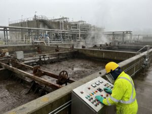 Arbeiter bedient Kontrollpanels an industrieller Kläranlage mit großem Absetzbecken und mechanischer Ausrüstung